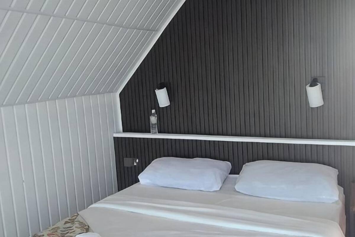 A bedroom with a sloped ceiling, a bed with white bedding, a bedside table, and a wall-mounted light fixture.