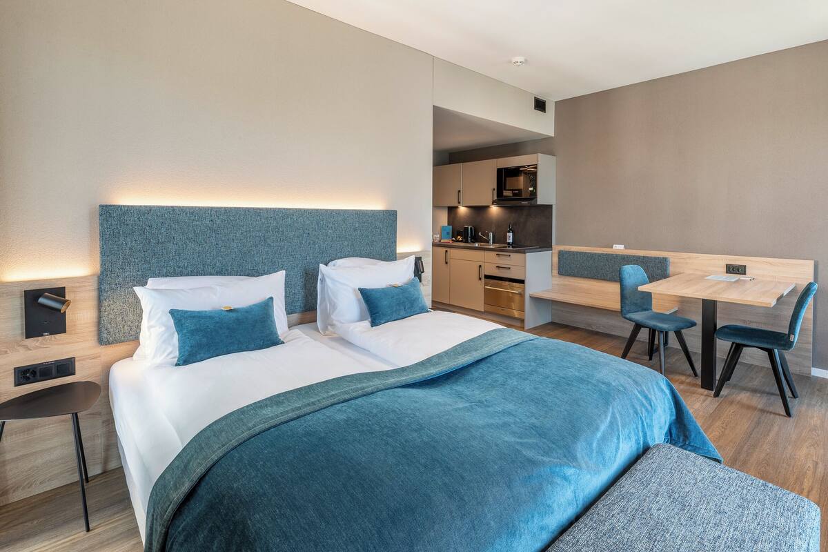 A modern hotel room with a large bed, a desk, and a kitchenette.