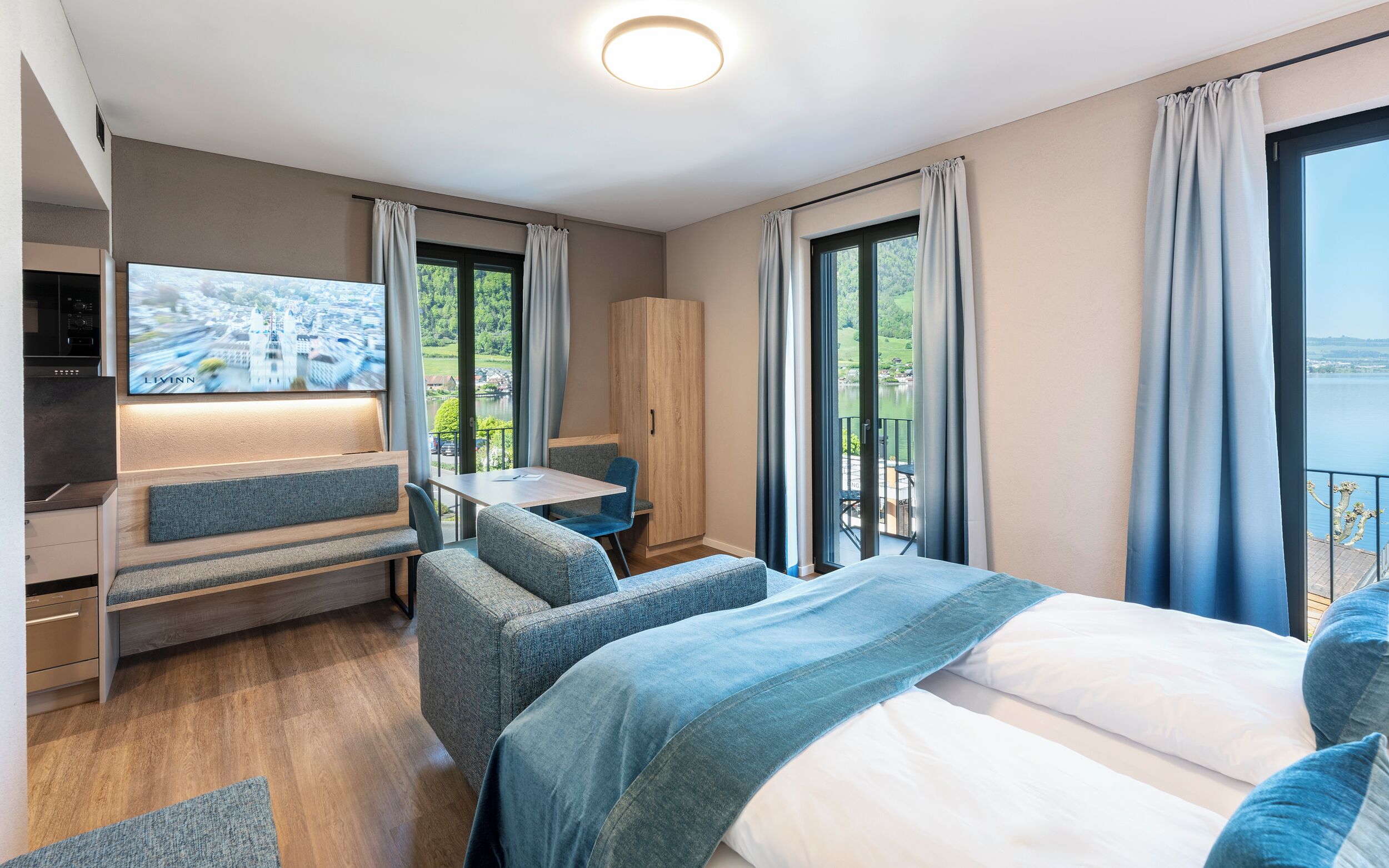 Una habitación de hotel moderna con una cama grande, un escritorio y un televisor. Cuenta con ventanas amplias con cortinas y un balcón con vista al lago.