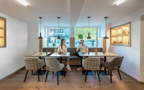 A modern dining area with a long table, four chairs, and a bench. There are pendant lights above the table, and a large window with a view of the street and greenery outside.