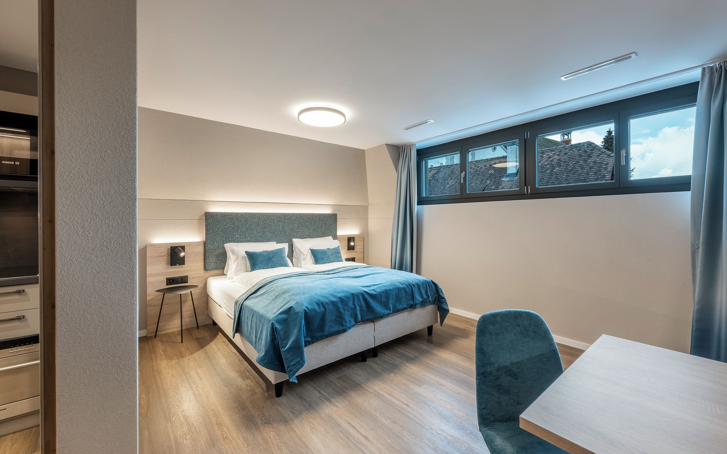 Une chambre moderne avec un lit, des tables de chevet, un bureau et une fenêtre donnant sur des immeubles.