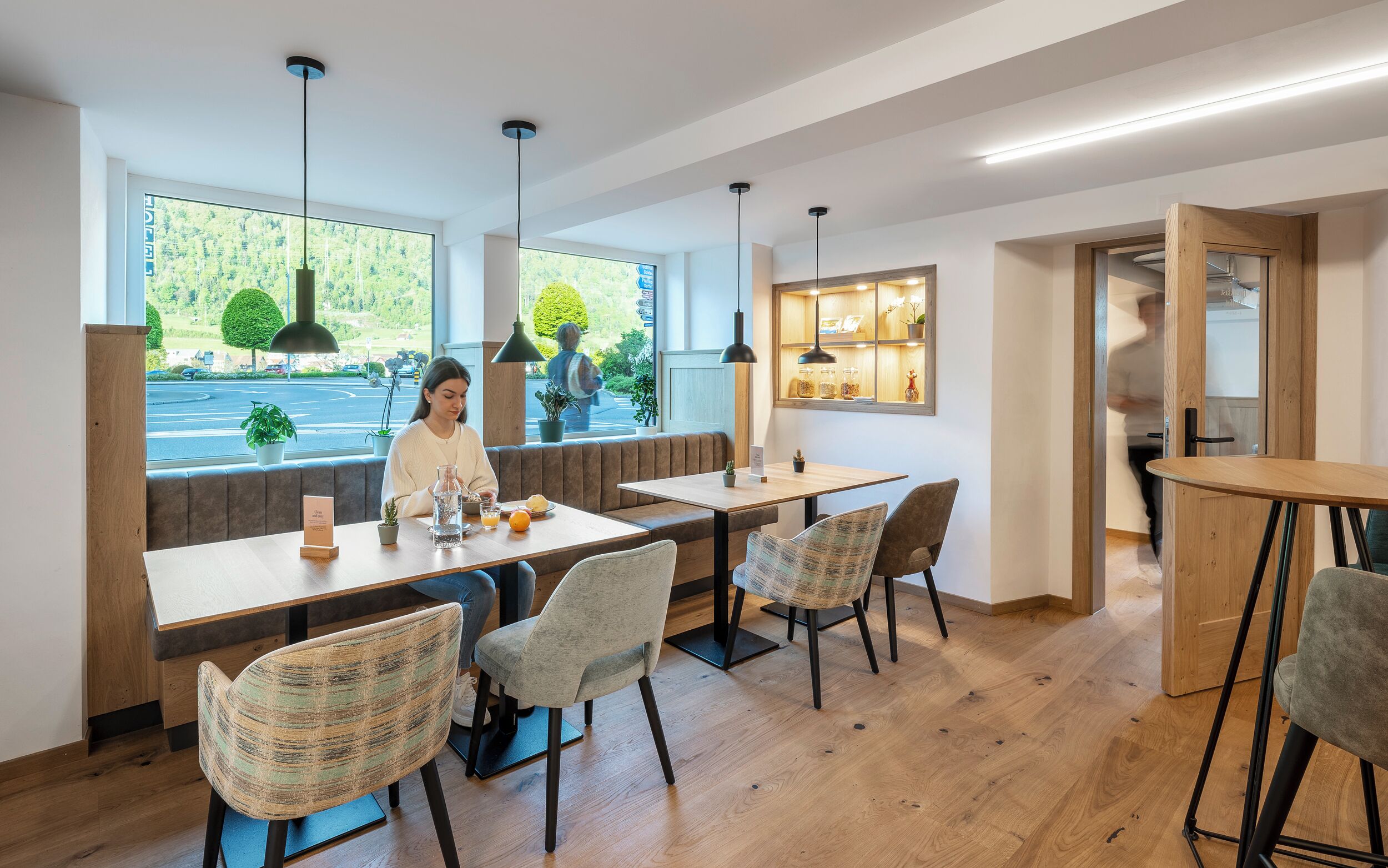 Un comedor moderno con mesas y sillas de madera, ventanas amplias con vistas a zonas verdes, iluminación colgante y una pared de madera decorativa con una vitrina.