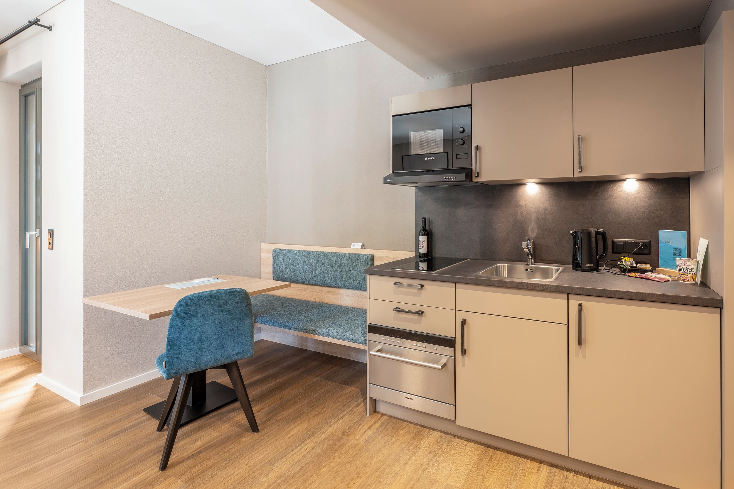 Cocina compacta con microondas, estufa y lavabo. Se observa una silla azul y una mesa pequeña.
