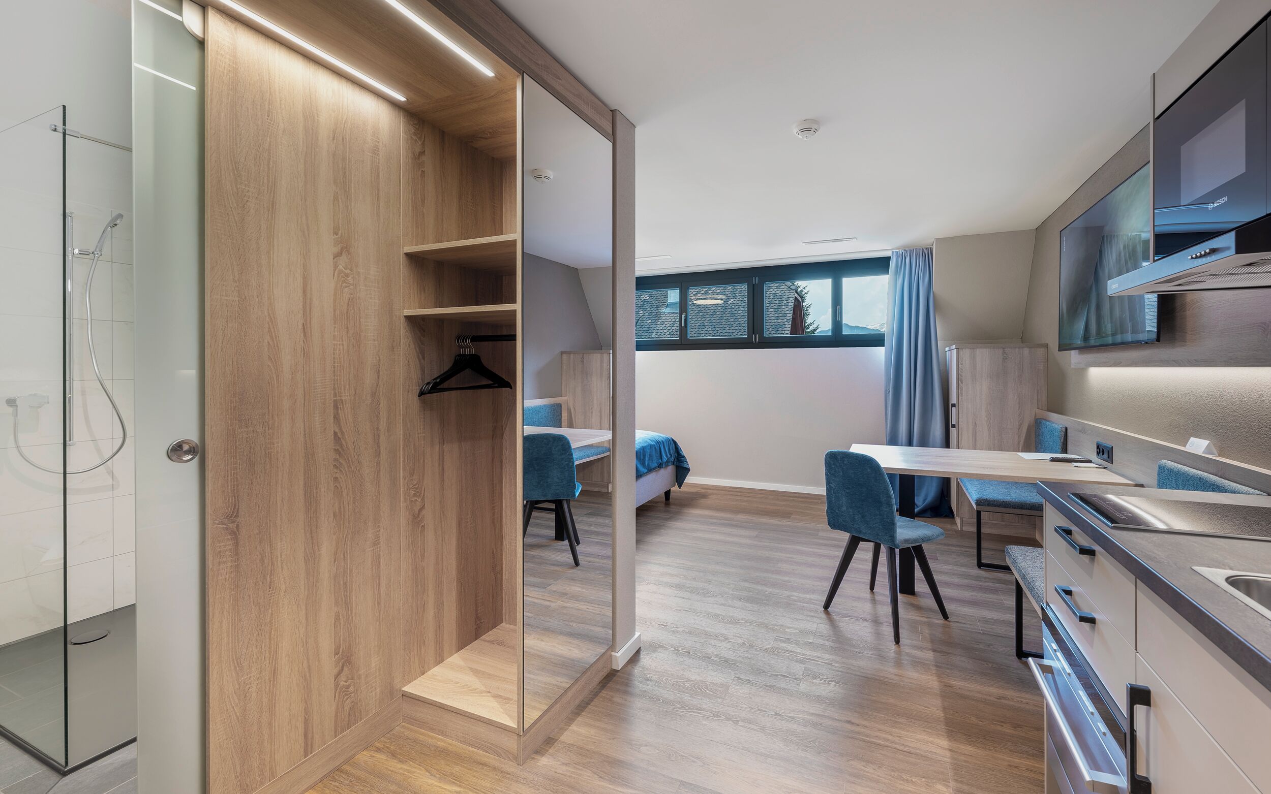 A modern kitchen with wooden cabinetry, a dining area with blue chairs, and a large window providing a view of the outdoors.
