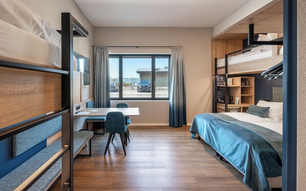 A compact hotel room with a bunk bed, a desk with a chair, and a large window offering a view of the outside.