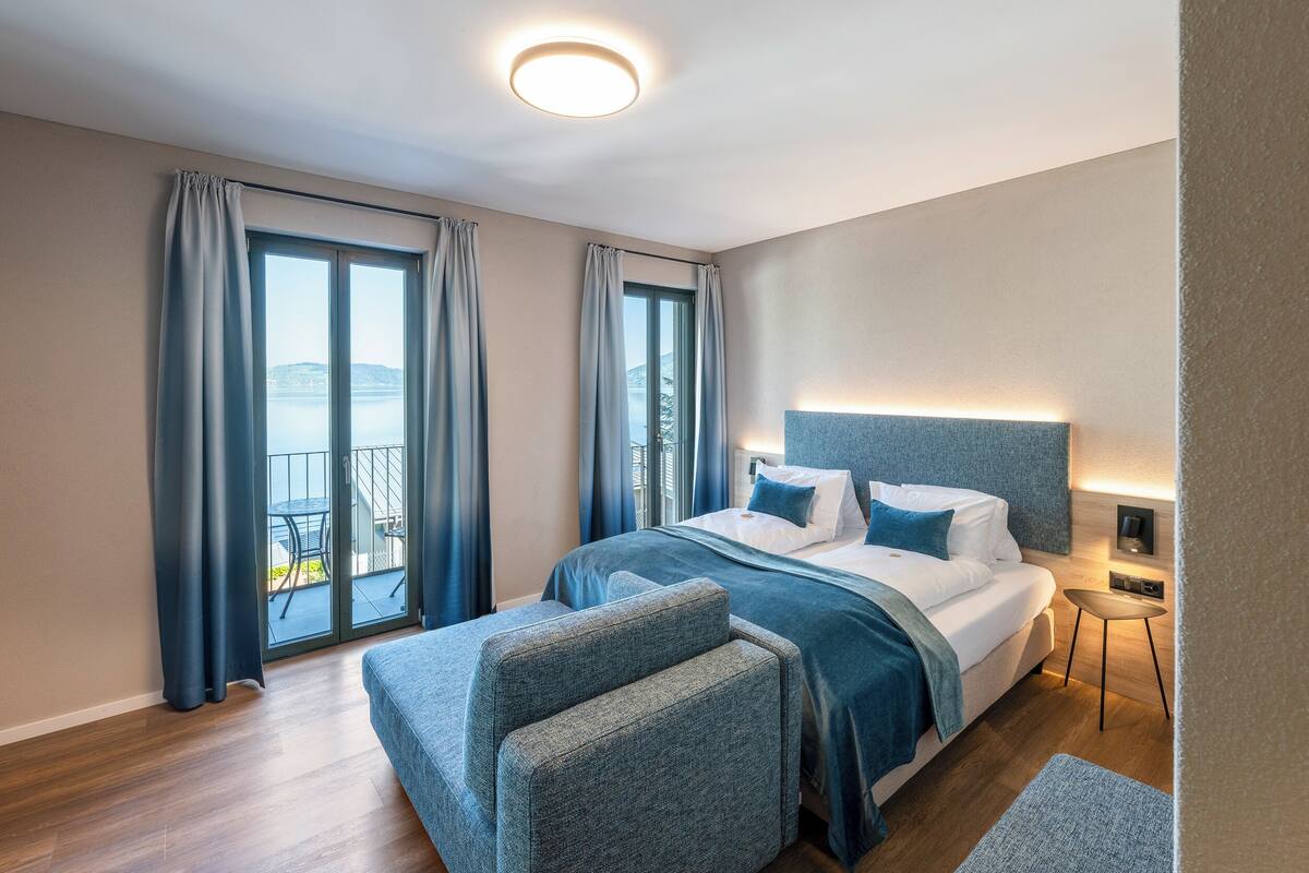 A modern hotel room with a large bed, a small side table, and a view of the sea through the windows.