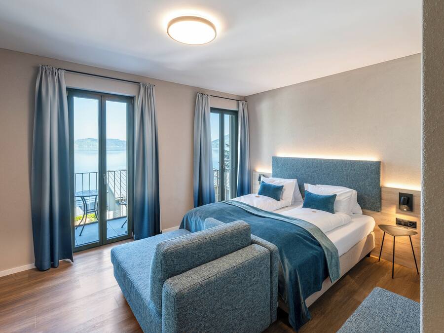 A modern hotel room with a large bed, a small side table, and a view of the sea through the windows.
