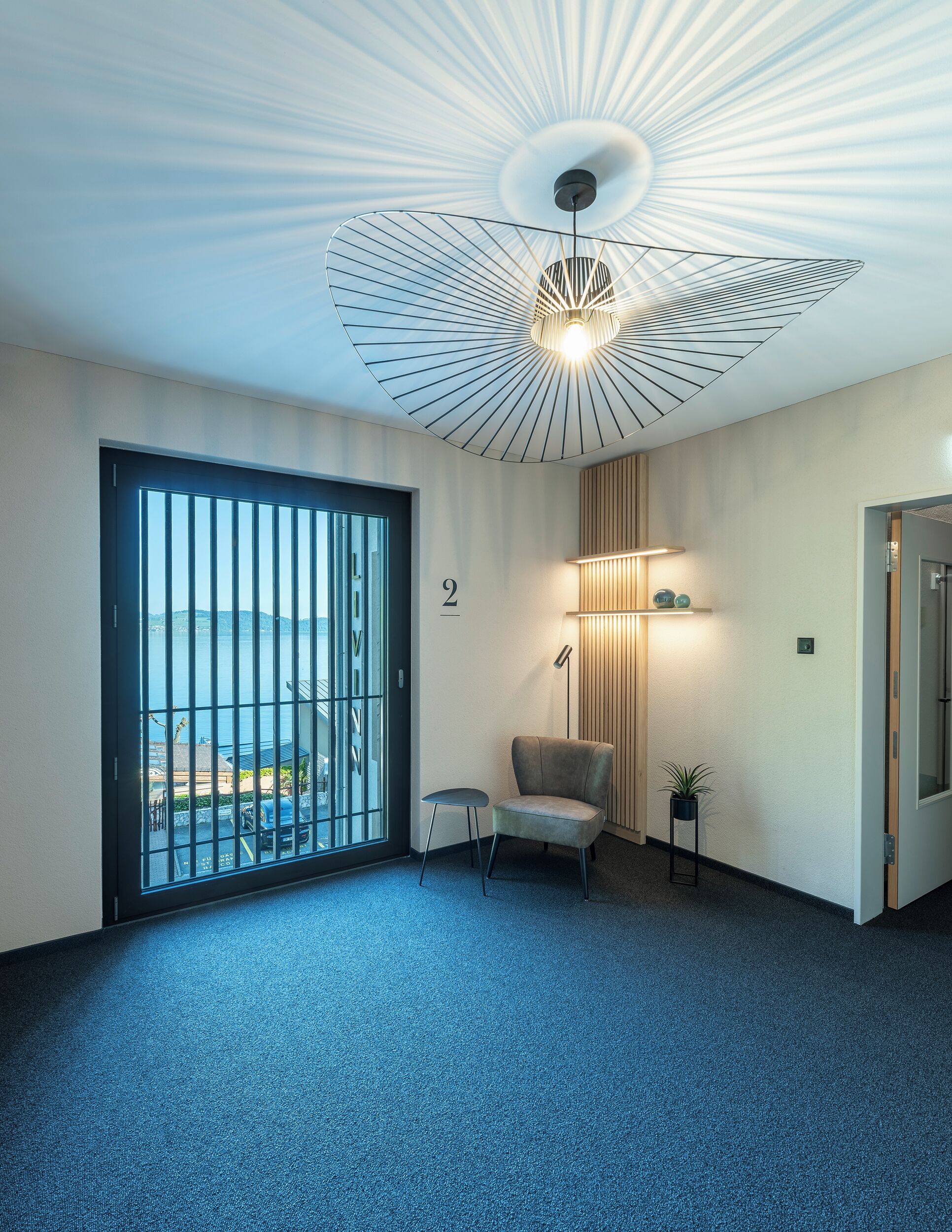 A modern office space with a large window, a unique ceiling light fixture, a small chair, and a potted plant.