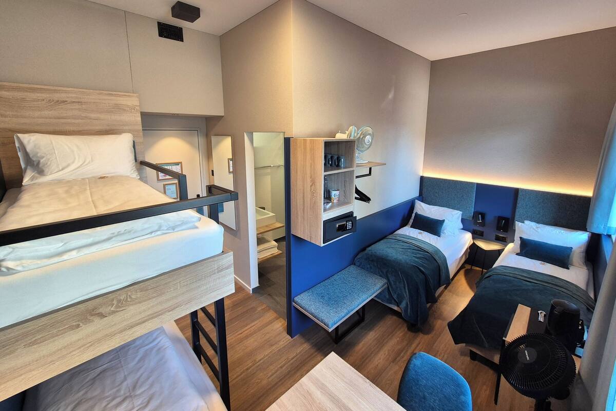 A modern hotel room with a bunk bed, a desk, a chair, a small table, a shelf with items, and a bedside table with a lamp.