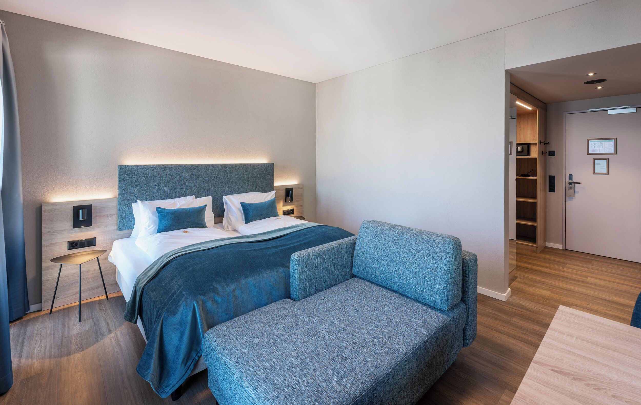 Ett modernt hotellrum med en stor säng, en blå stoppad fåtölj, ett litet runt bord och en inbyggd garderob.