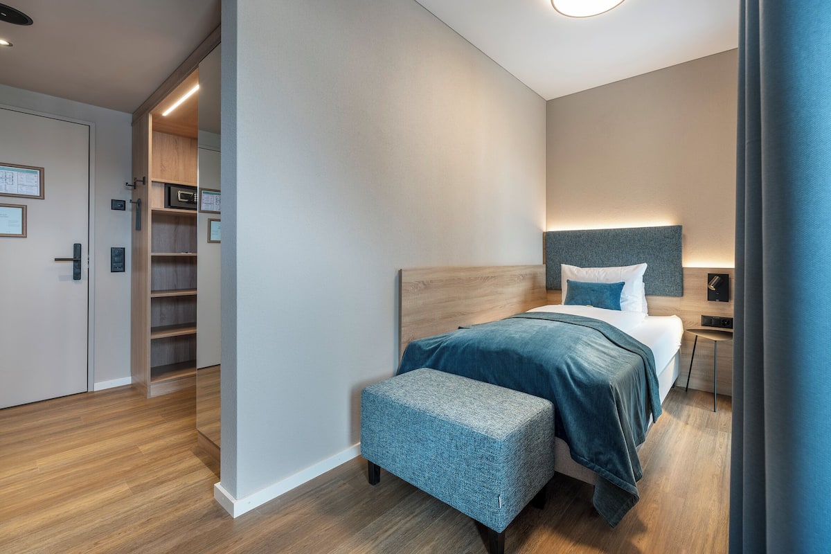 A modern hotel room with a wooden headboard, a bed with a blue blanket, a bedside table, and a small bench.