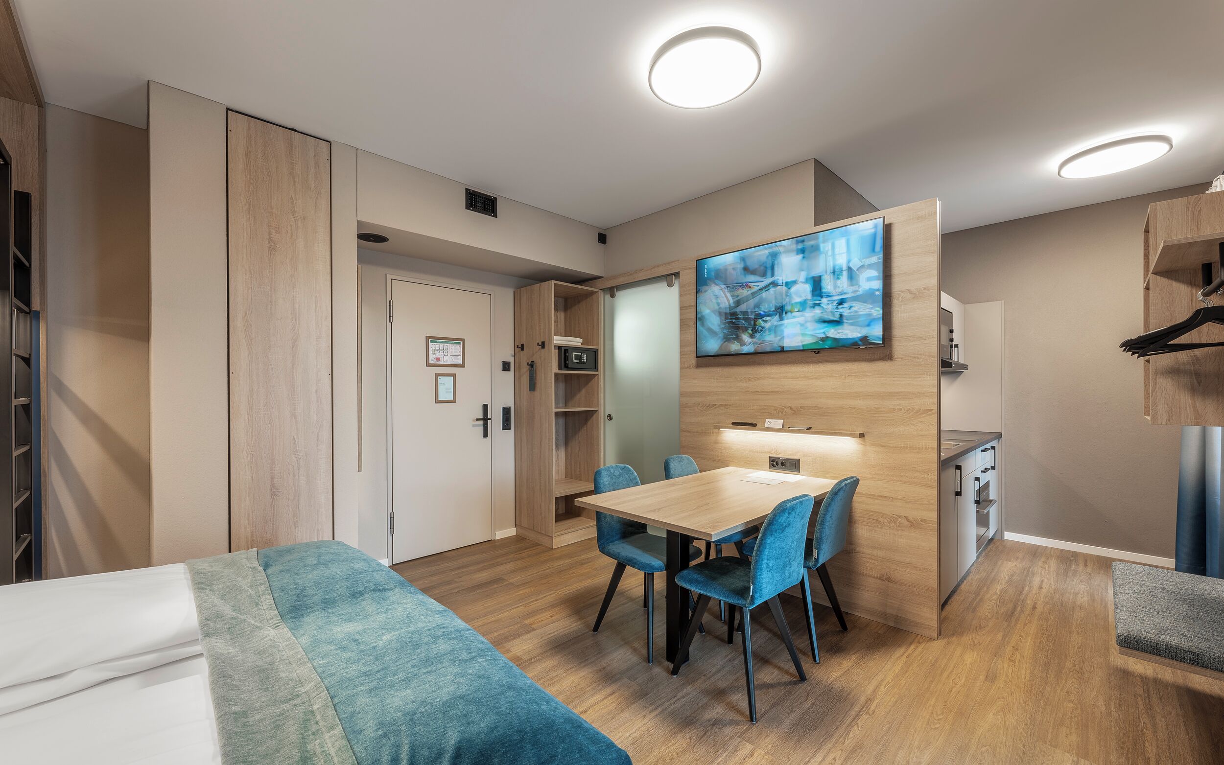 Una habitación de hotel moderna con una mesa de comedor de madera, sillas azules, un televisor de pantalla plana grande y una kitchenette.