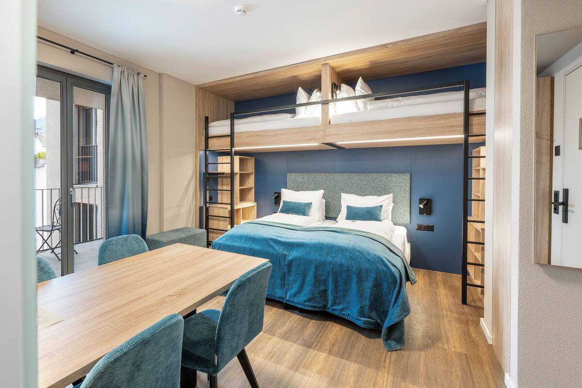 A compact hotel room with a bunk bed, a dining table with chairs, and a small seating area by the window.