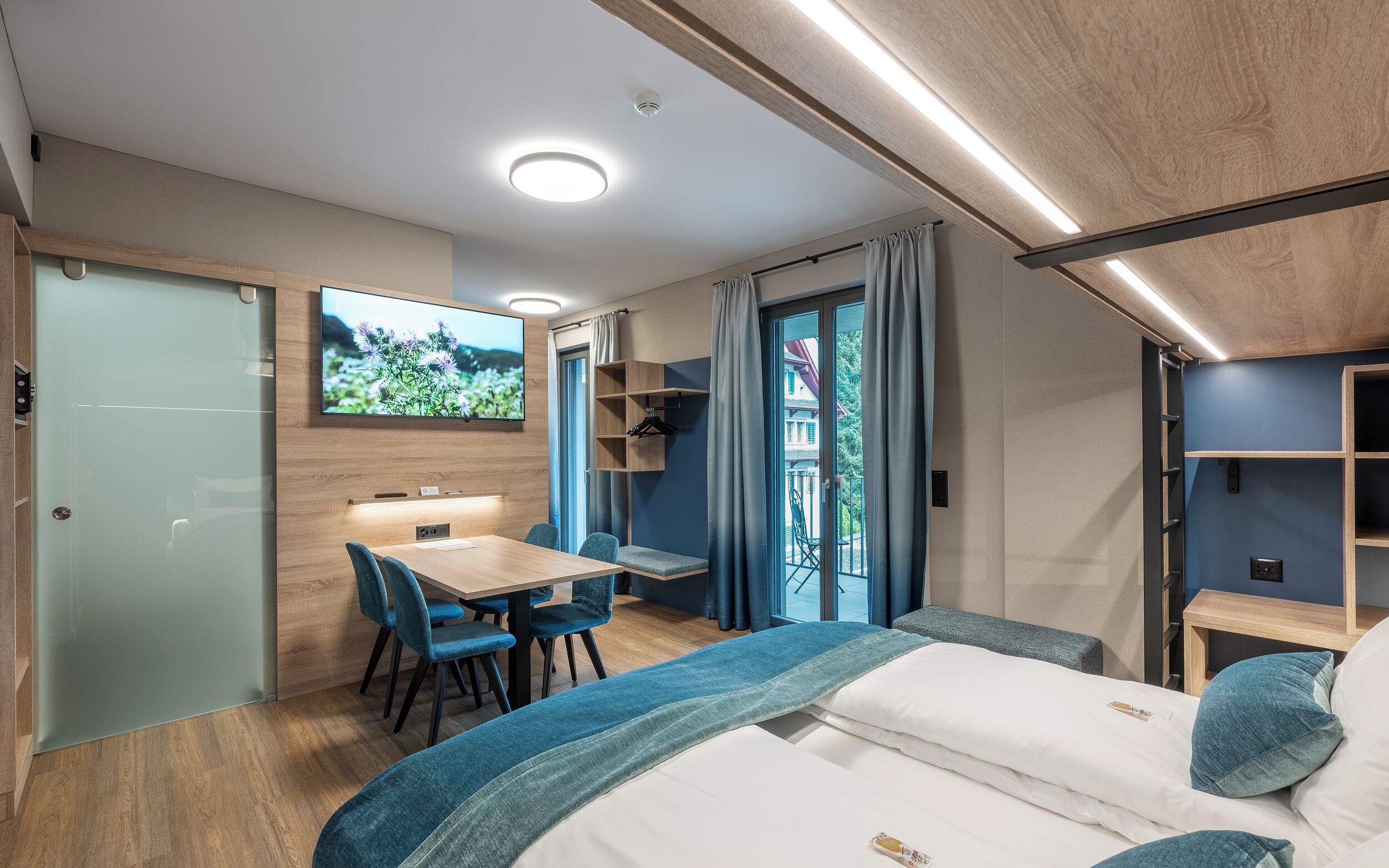 A compact room with a bed, a desk, and a chair. There is a television mounted on the wall, and a window with curtains.