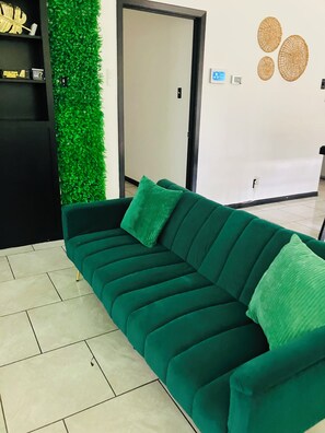 A green sofa with two cushions, a black shelving unit with decorative items, a green wall with a textured pattern, and a white door in the background. - The Greenhouse Effect- central+ 4BR/2BA+ king suite+ coffee+ BBQ grill (Corpus Christi)