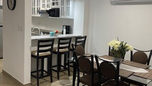 A modern kitchen with white cabinets, a microwave, and a coffee maker. There is a dining area with a table and chairs, and a clock on the wall.