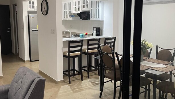 A modern kitchen with white cabinets, a microwave, and a refrigerator. There is a dining area with a table and chairs, and a gray sofa in the living room.