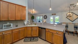 A kitchen with wooden cabinets, a dishwasher, a toaster, pendant lights, and a dining area with chairs and a table. - Sunset Serenity in Port Charlotte (Port Charlotte)