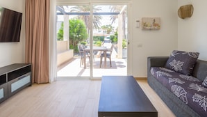 Ein Wohnzimmer mit einer Couch, einem Fernseher, einem Couchtisch und einer Glastür, die auf eine Terrassenanlage mit Tisch und Stühlen führt.