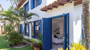 A white building with blue doors and windows, a white couch inside, and a small garden with plants. - House in Búzios less than 100m from the beach (Vila Luiza Armação dos Búzios)