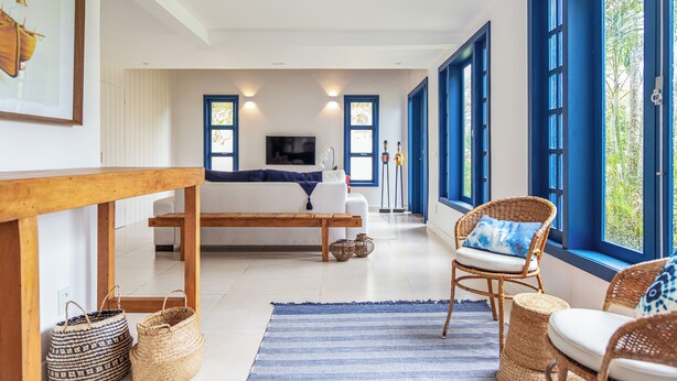 A spacious living room with a wooden bench, wicker chairs, a large window with blue shutters, and a flat-screen TV mounted on the wall. - House in Búzios less than 100m from the beach (Vila Luiza Armação dos Búzios)
