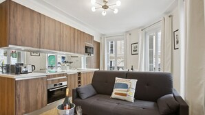 Eine moderne Küche mit integrierter Kochinsel, ein graues Sofa mit Zierkissen und ein Esstisch mit Obstschale und Champagnerflasche.