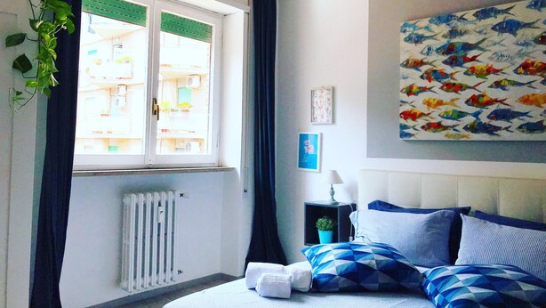 A hotel room with a bed, a window with curtains, a radiator, a nightstand with a lamp, and a wall with a fish painting. - Cozy Vintage Apartment in Trastevere within walking distance of downtown  (Roma)