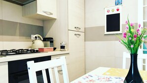A kitchen with a gas stove, a kettle, and a coffee maker. There is a white chair and a table with a floral tablecloth. A vase with flowers is on the table. - Cozy Vintage Apartment in Trastevere within walking distance of downtown  (Roma)