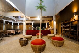Un restaurante con techo alto, vigas a la vista y un área de bar central. Hay sillas de mimbre con cojines rojos y un pequeño árbol en el centro.