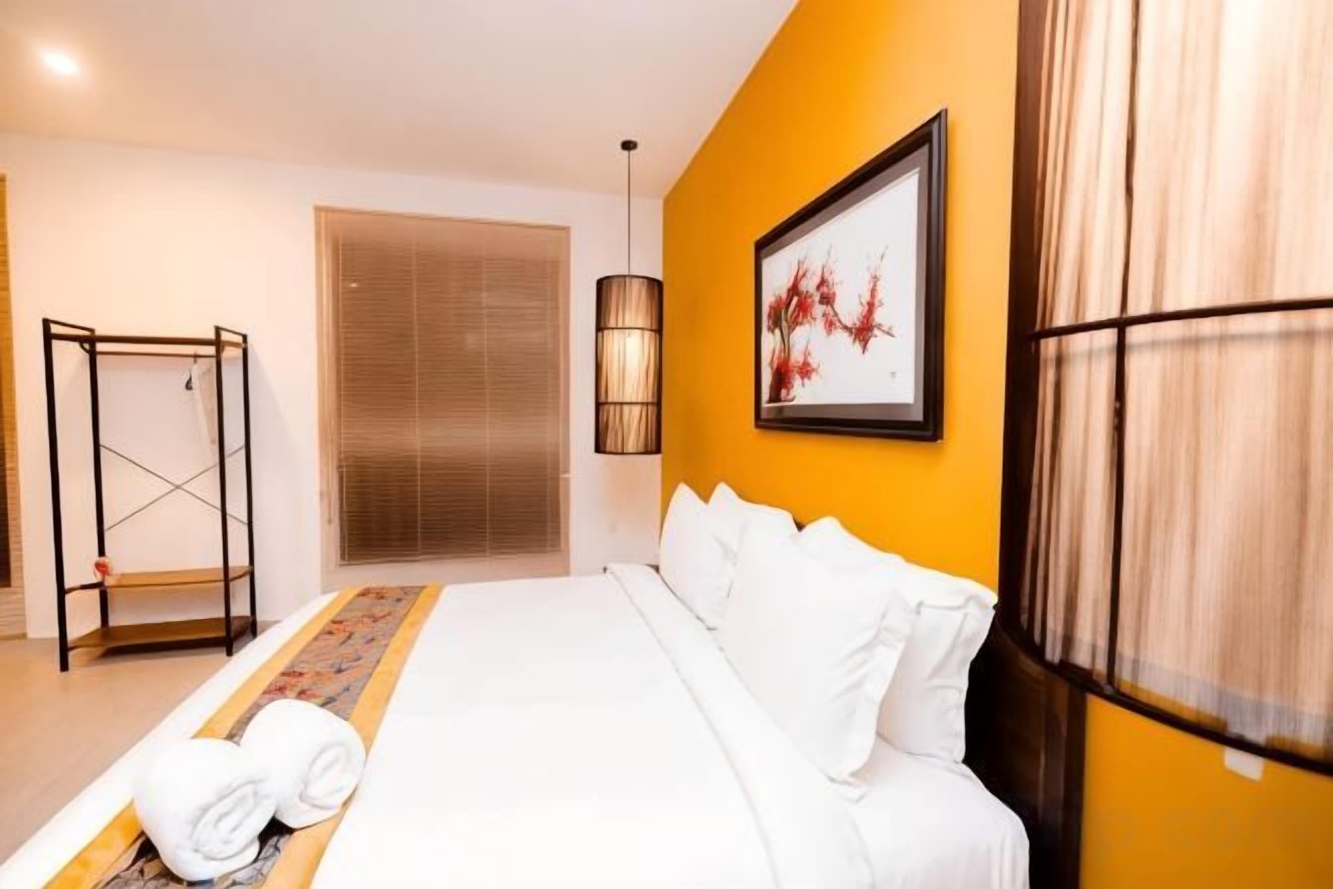 A hotel room with a bed, a window with blinds, a wall-mounted artwork, and a standing lamp.