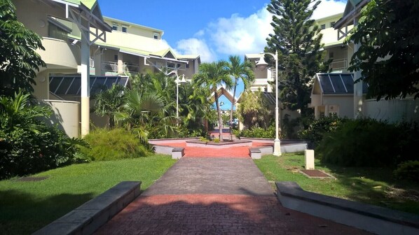 A pathway leading to a building with a red brick entrance, surrounded by greenery and palm trees. - T3 seaside duplex (Pointe-à-Pitre)