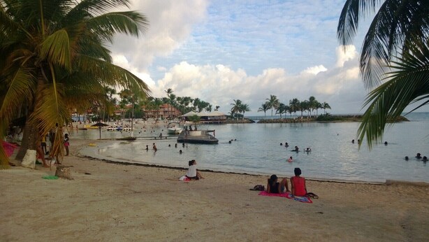 Ein Strand mit badenden und entspannten Menschen. Palmen und ein Boot liegen im Wasser.