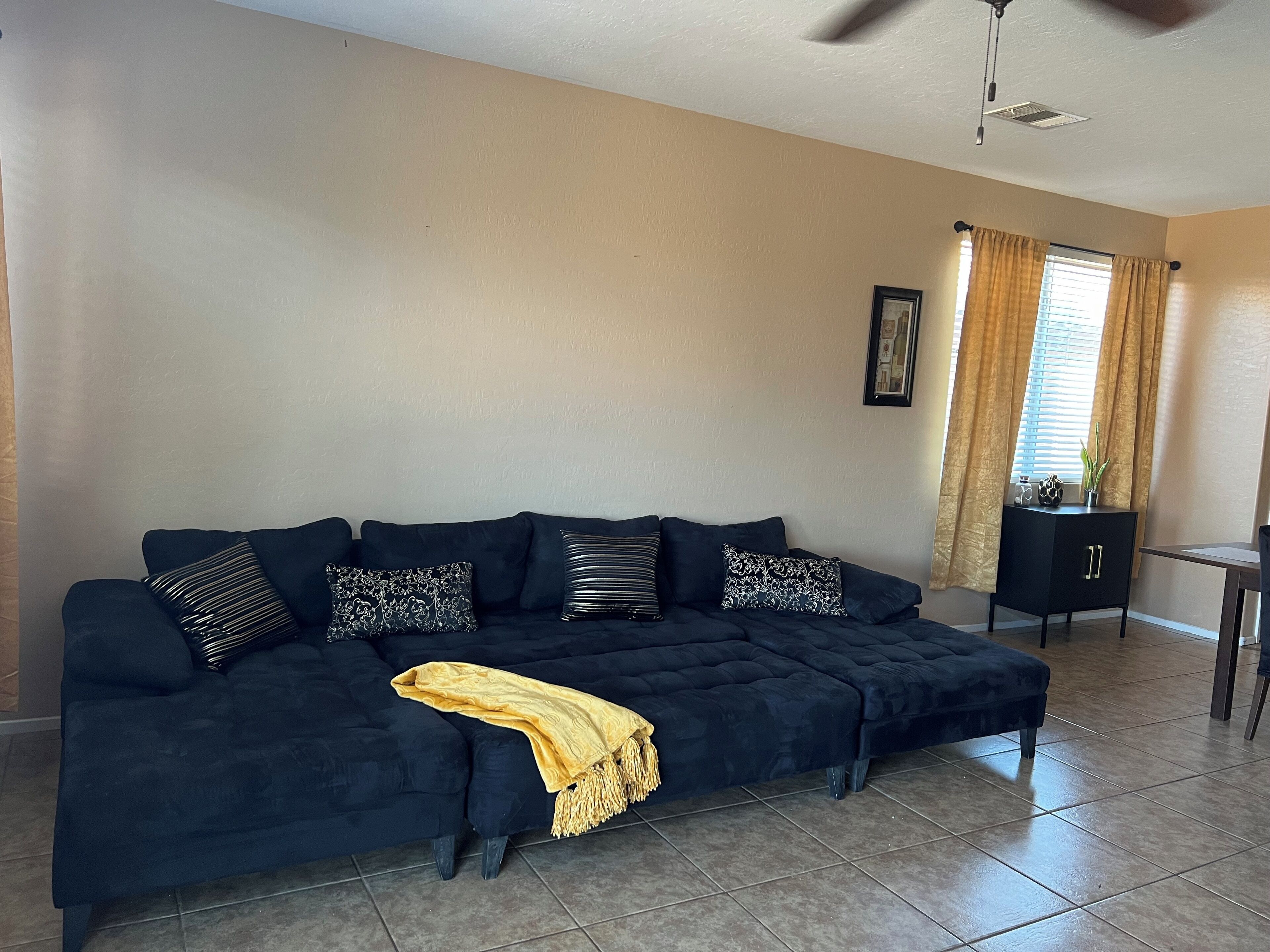 A dark blue sofa with patterned cushions, a yellow throw blanket, and a ceiling fan in a room with beige walls and a tiled floor.