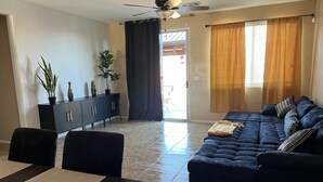 Una sala de estar con un sofá azul oscuro, una mesa de comedor con sillas, un mueble negro con objetos decorativos y un ventilador de techo.