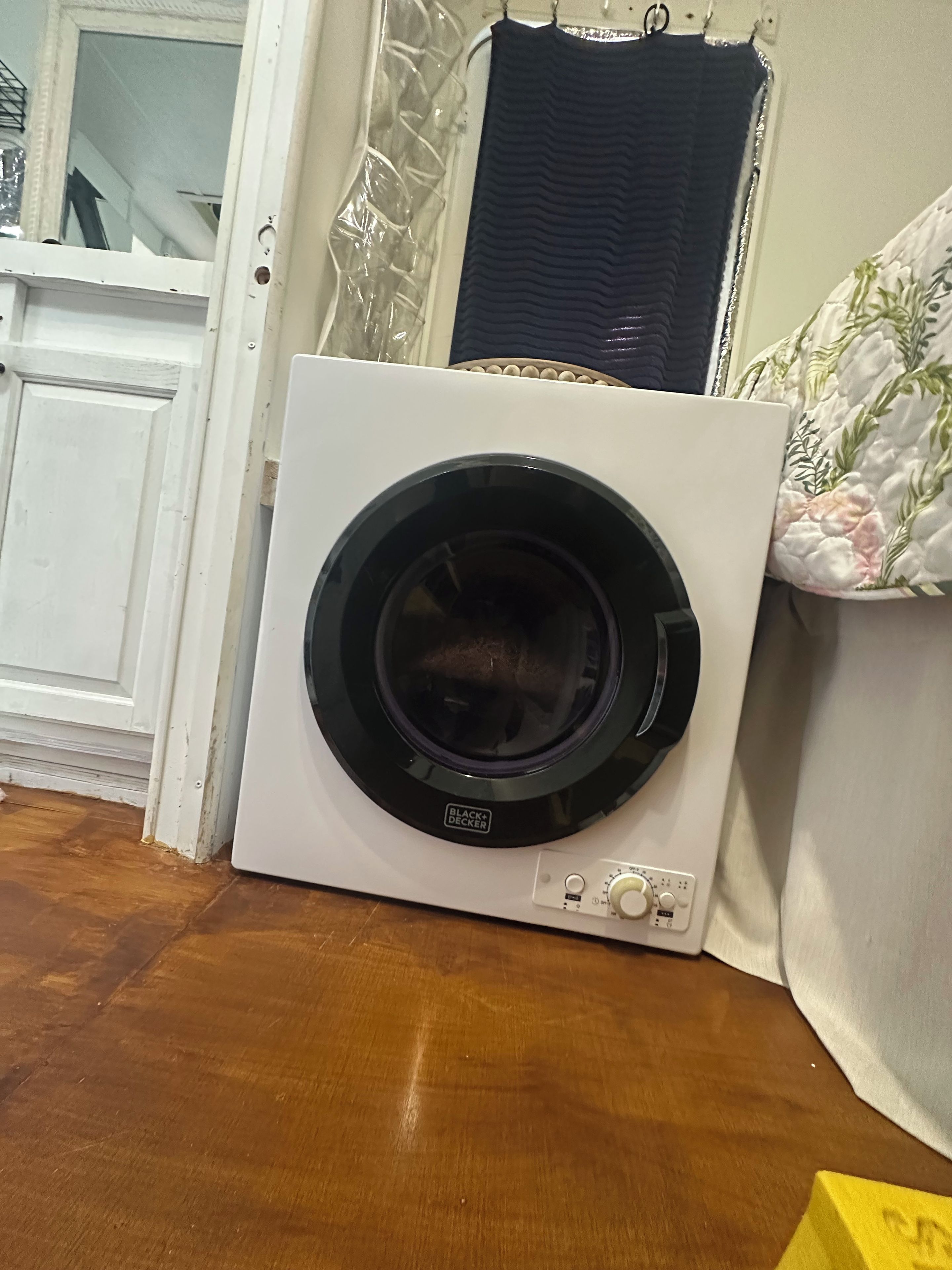 A front-loading washing machine with a control panel, placed on a wooden floor next to a white door and a window with a curtain.