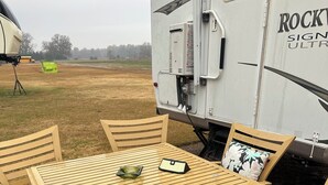Ein Holztisch mit Stühlen steht auf einer Ziegelterrasse, im Hintergrund ist ein Anhänger zu sehen.