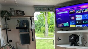 Ein Zimmer mit Fernseher, der eine Streaming-Dienstanwendung anzeigt, einem kleinen Tisch mit Fernbedienung, einer gepolsterten Bank und Blick auf eine grüne Rasenfläche durch eine Tür.