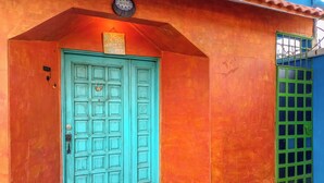 Een turquoise deur met een kleine opstap, erboven een wandklok en een raam met tralies.