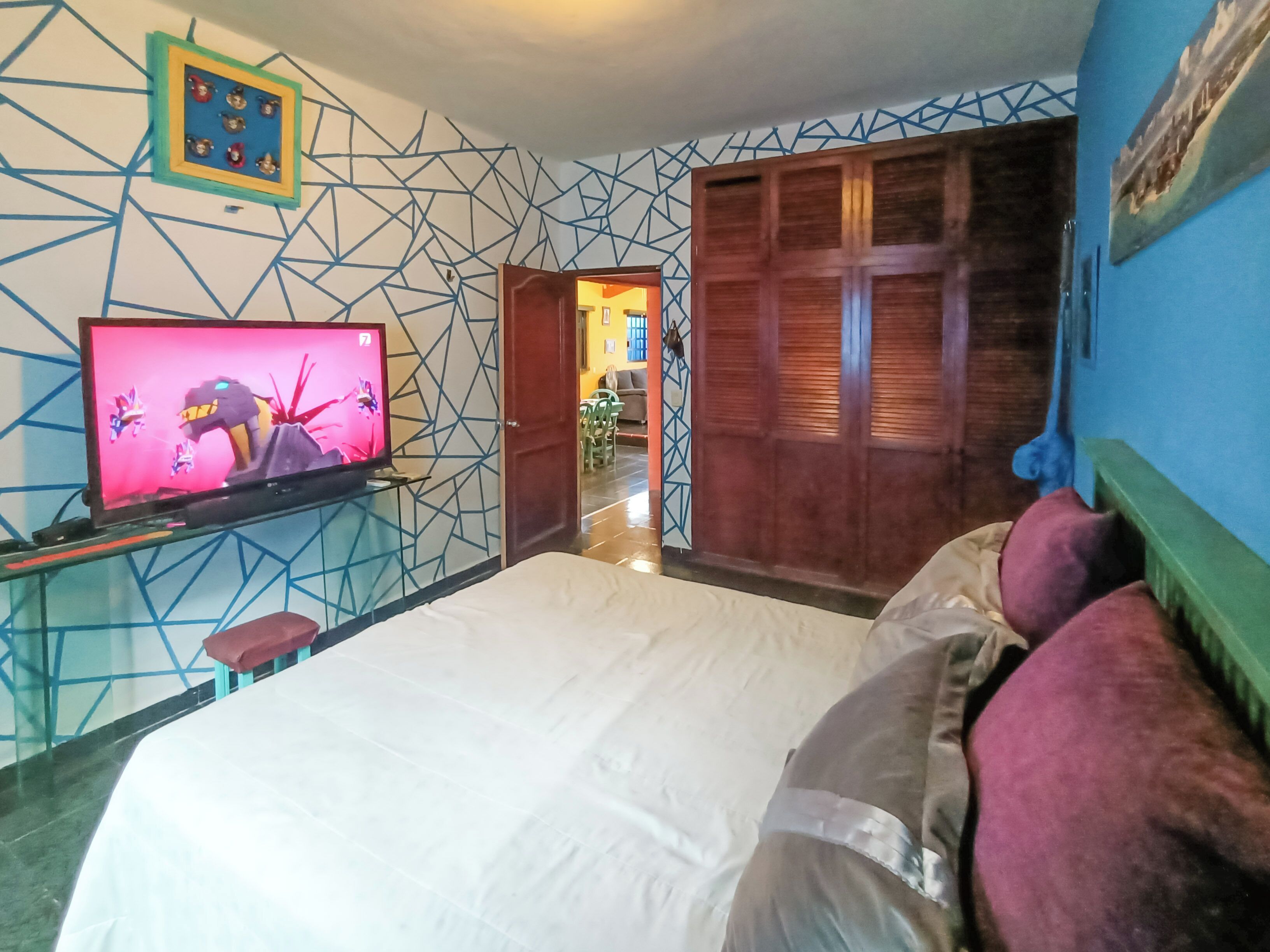 A hotel room with a bed, a television displaying a cartoon, a geometric-patterned wall, and a wooden wardrobe.