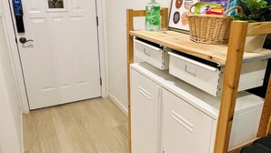 Una habitación compacta con una puerta blanca, un pequeño estante con una planta, una cesta de mimbre y un cuadro en la pared.