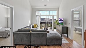 Ein modernes Wohnzimmer mit einem grauen Ecksofa, einem Deckenventilator, einem Fernseher und einem Balkon mit Blick auf die Stadt.