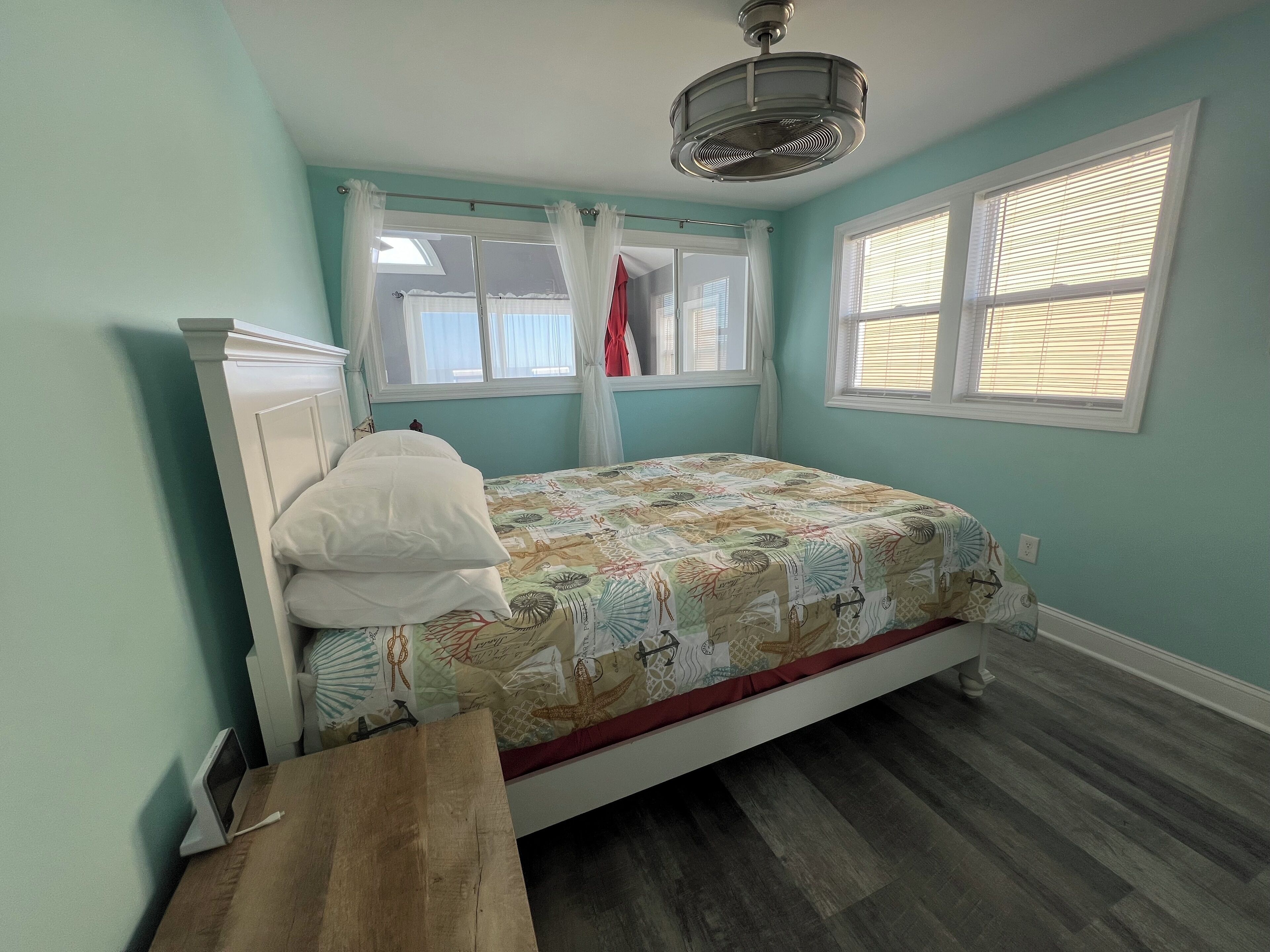 A bedroom with a bed, a bedside table, a ceiling fan, and two windows with curtains.