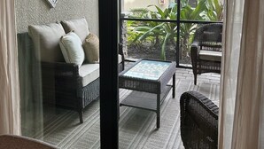 Een kamer met een glazen deur die uitkomt op een patio. Er staat een rieten bank met kussens, een kleine tafel en een rieten fauteuil. De patio heeft een betegelde vloer en is omringd door planten en een gebouw met een pannendak.