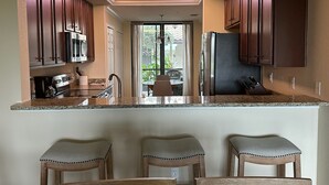 Een keuken met donkere houten kastjes, een roestvrijstalen koelkast, een magnetron en een granieten aanrecht. Drie barkrukken en een eettafel met stoelen zijn zichtbaar.