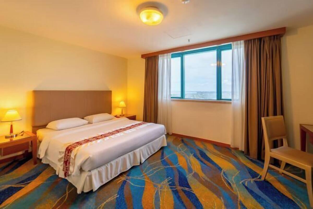 Habitación de hotel con cama, lámparas de noche, una silla, ventana con cortinas y vistas de la ciudad.
