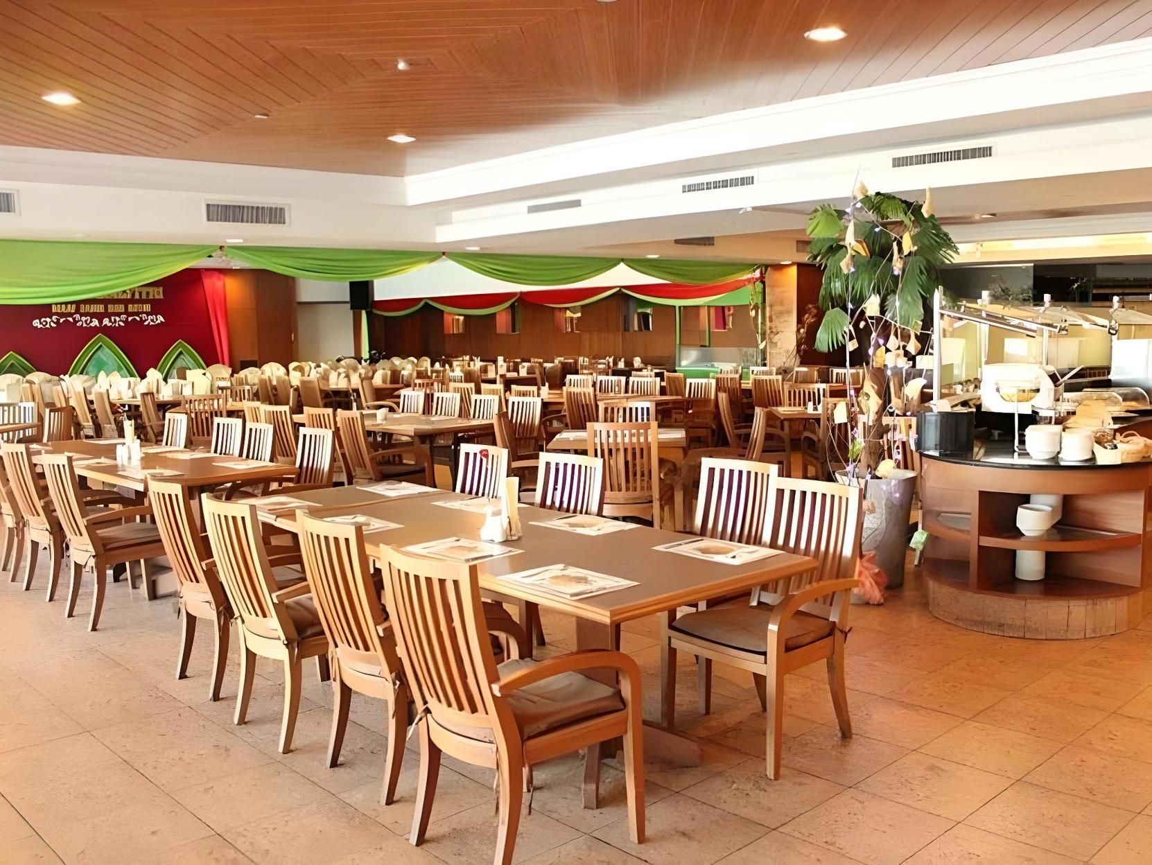 A spacious indoor dining area with multiple wooden tables and chairs, a kitchen area with cooking equipment, and a decorative plant arrangement.