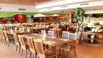 A spacious indoor dining area with multiple wooden tables and chairs, a kitchen area with cooking equipment, and a decorative plant arrangement.