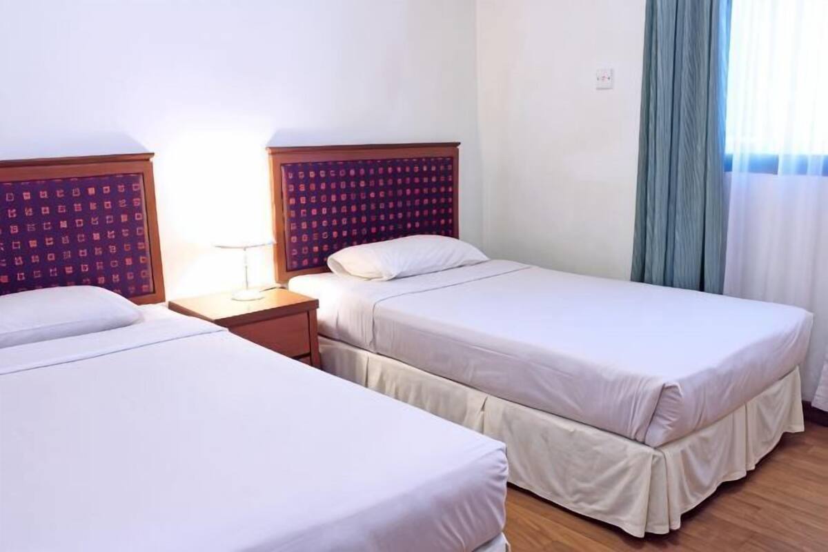 Habitación de hotel con dos camas individuales, una mesita de noche con lámpara y una ventana con cortinas.