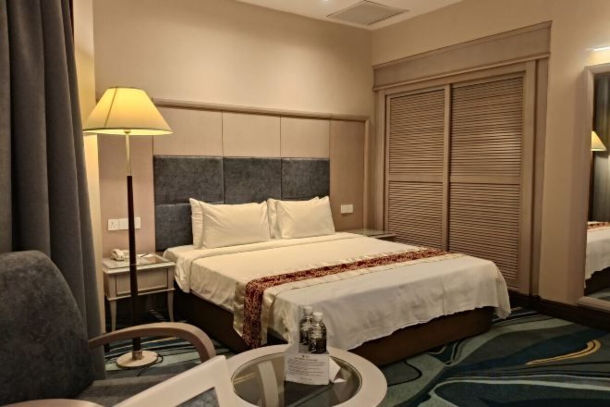 A hotel room with a bed, bedside table, lamp, and a view of the outside.