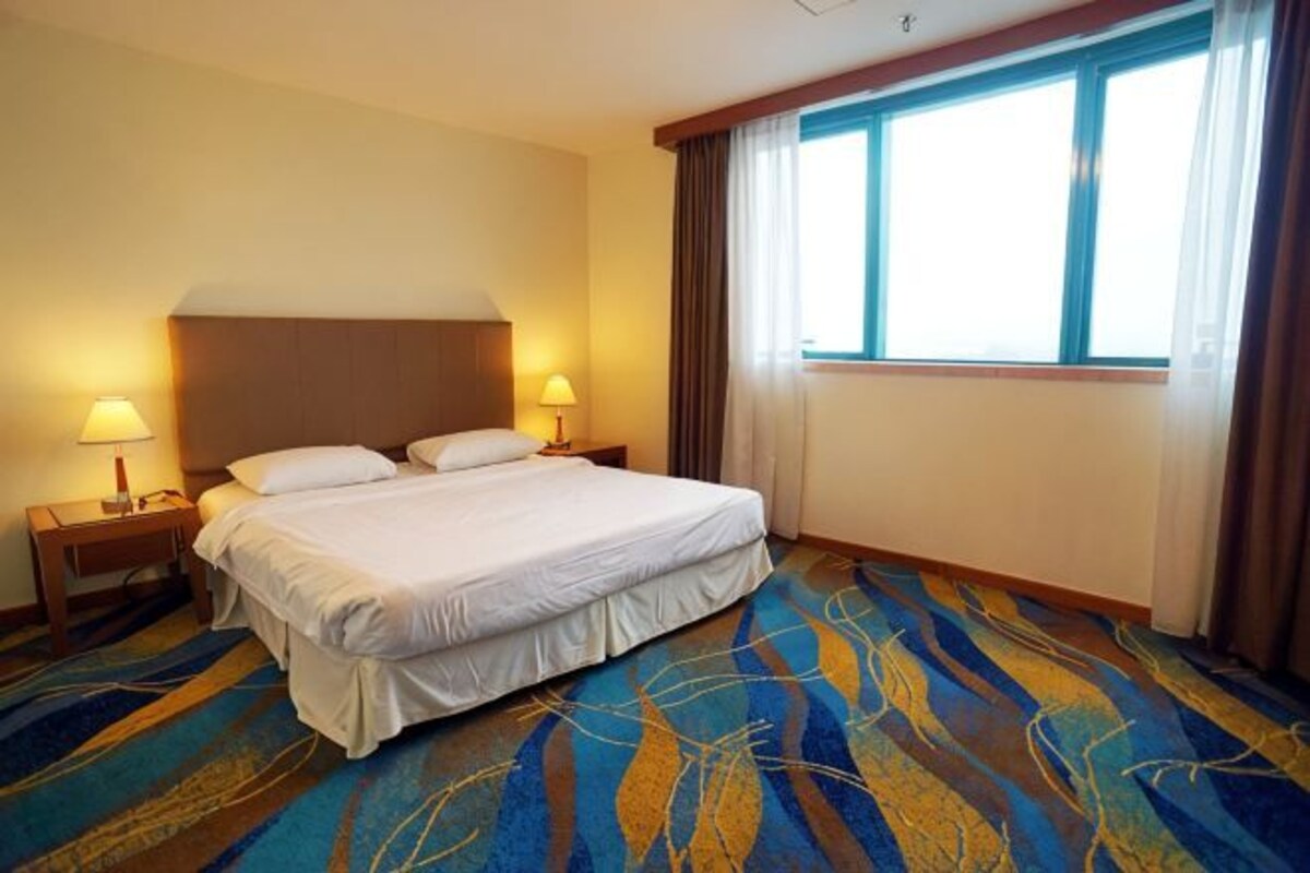 A hotel room with a large bed, two bedside lamps, a window with curtains, and a patterned carpet.